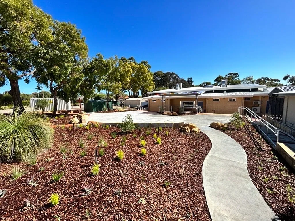 Curved grey concrete path running out to a larger paved area past new planting at Majella Primary School